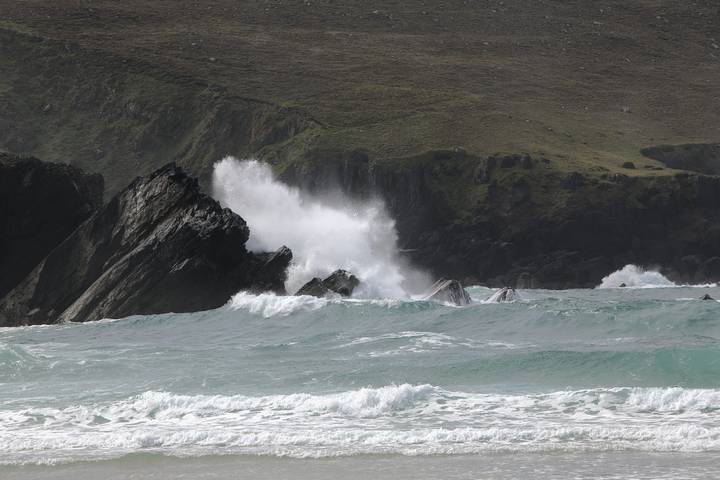 Rough turquoise waves crashing violently against dark jagged rocks along a rugged coastline.