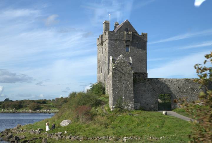 Medieval stone castle perched beside a river with blue skies and grassy banks.