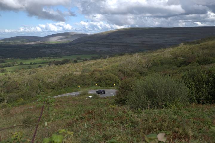 Curving rural road descending through green hills with a car navigating the bend below dramatic skies.