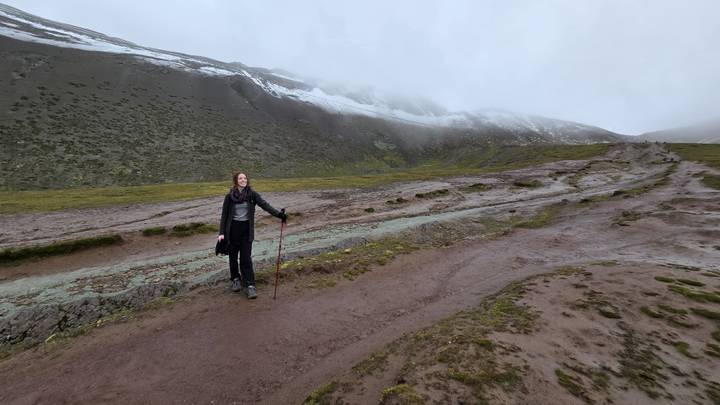 A lone hiker with a trekking pole stands on a misty Andean mountain trail with patches of snow on the slopes around.