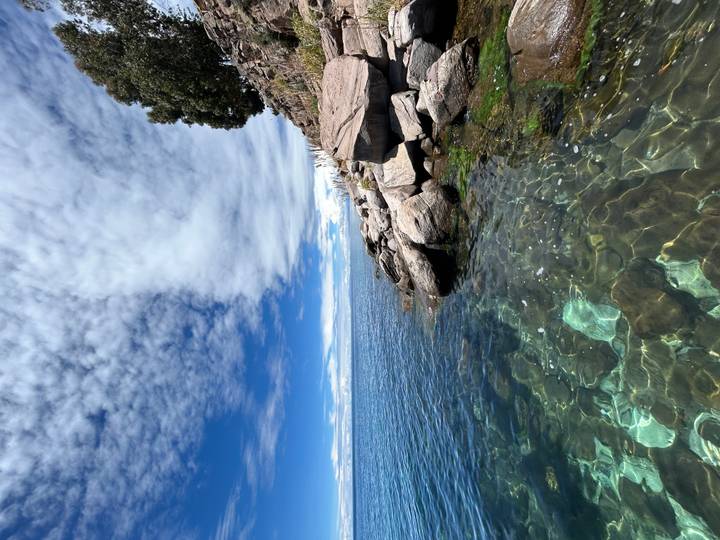 Rocky shoreline with crystal-clear turquoise water and a dramatic sky over a vast high-altitude lake.