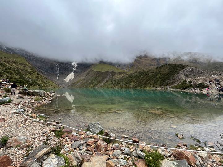 Emerald-green alpine lake beneath steep, cloud-shrouded peaks with small groups of visitors along the shore.