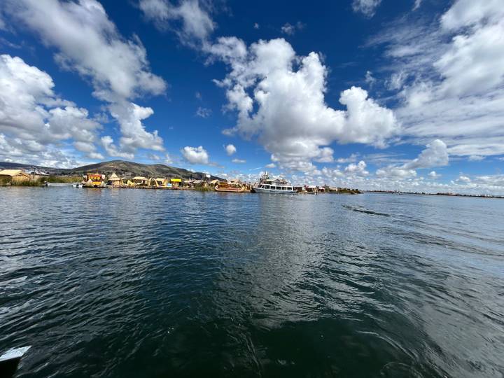 Expansive lake with floating reed islands and boats under a sky dotted with bright white clouds.