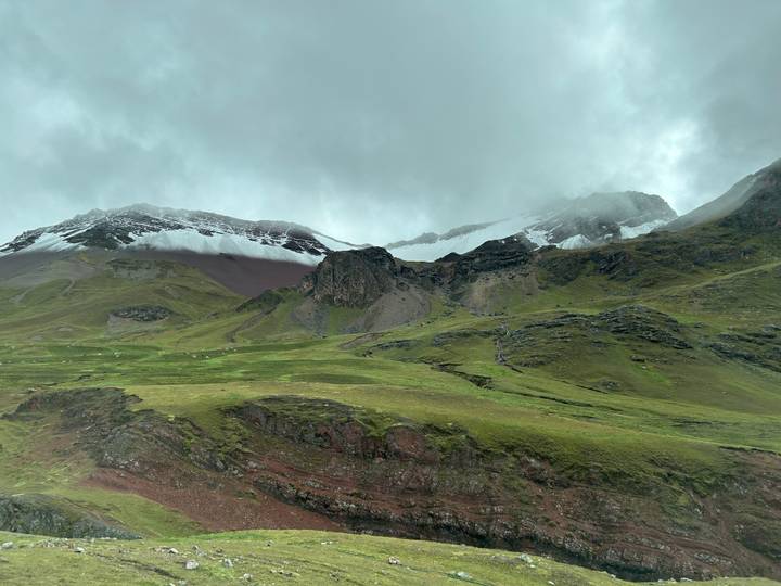 Green pastures and red earth rise to snow-streaked peaks hidden by low clouds.