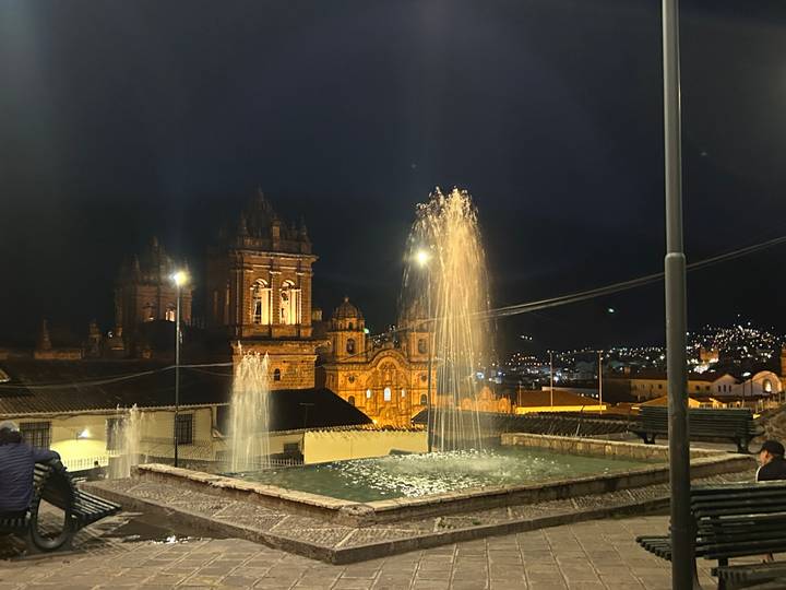 Night scene of Cusco cathedral and fountain jets glowing under warm lights.