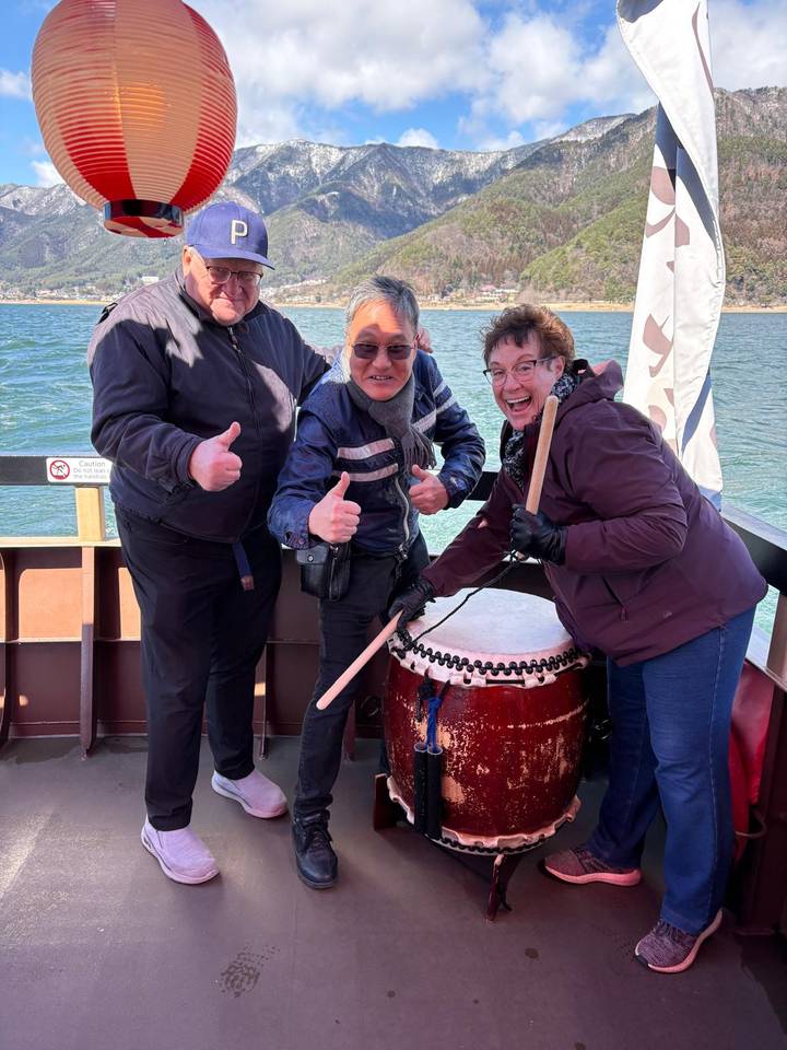 Smiling travelers giving thumbs up while trying a traditional drum on a boat over a blue lake.