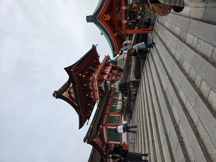 Vermilion-and-white shrine building atop stone steps with visitors ascending under cloudy skies.
