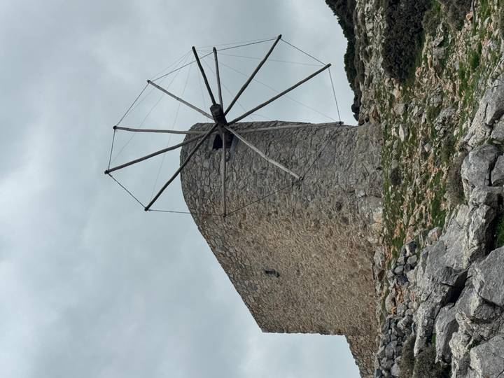 Stone windmill ruin with wooden sails perched on a rocky hillside under cloudy skies.