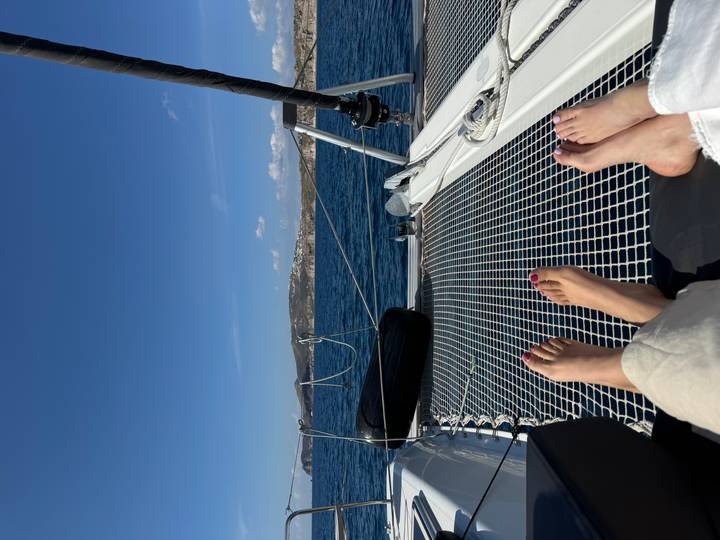 Two people relax with their feet on the netted bow of a catamaran sailing the Aegean.