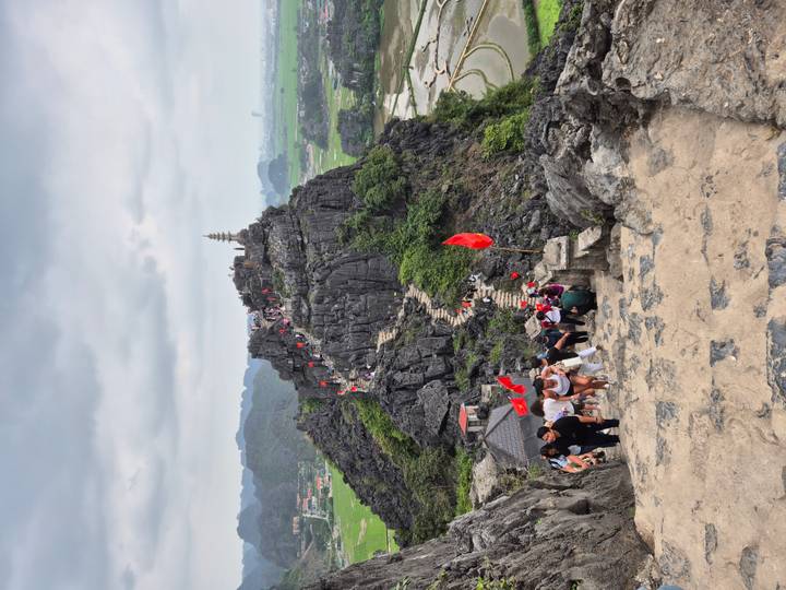 Steep stone stairway leading up a jagged limestone peak with red flags and many hikers visible, overlooking verdant rice fields.