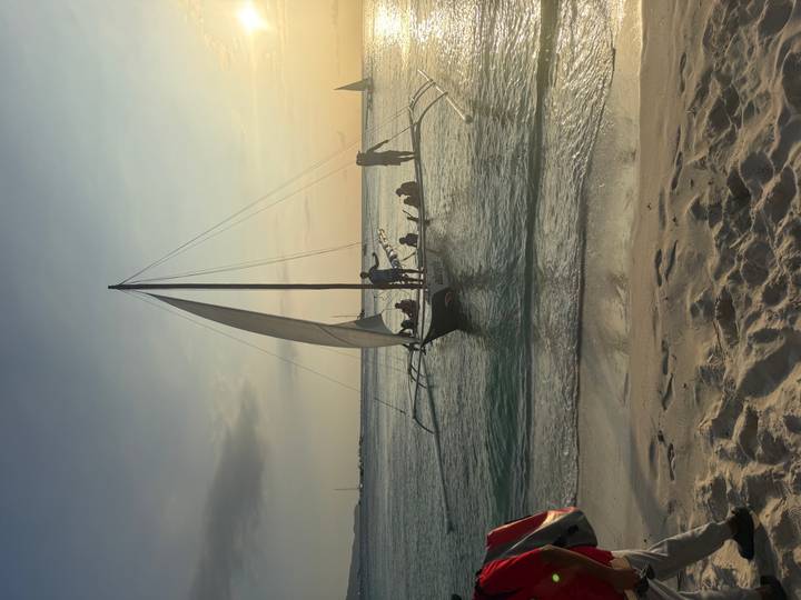 Sailboat with lateen sail carries passengers on calm turquoise water at sunset near the shore.