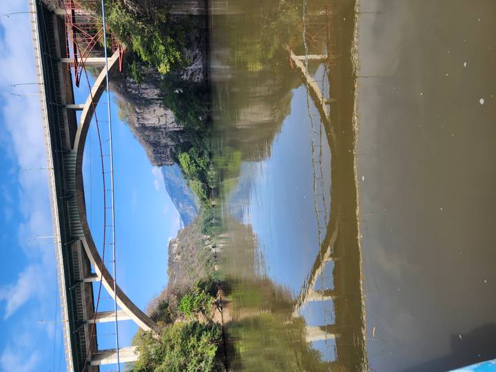 Tall bridge spanning a calm river with a reflection of the arch, framed by steep green cliffs and distant mountains.