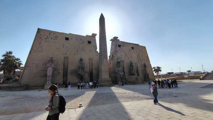 Ancient temple entrance with towering obelisk backlit by the sun, visitors gathered around.
