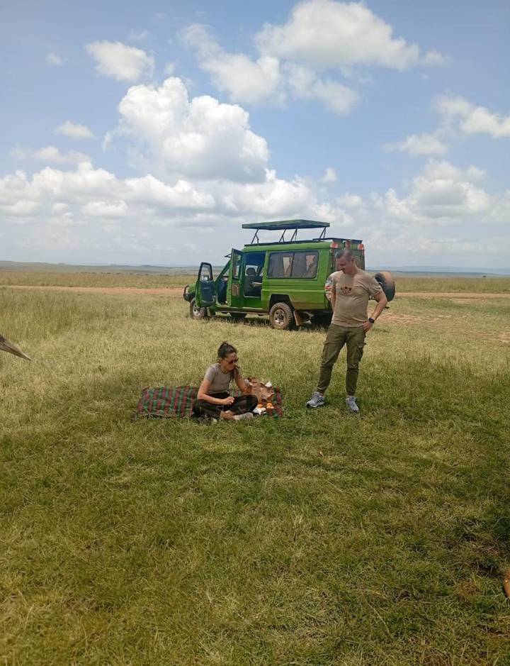 Two safari travelers enjoying a picnic beside a green 4x4 on the savannah.
