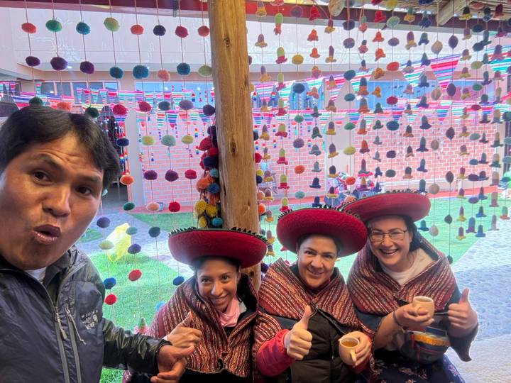 Group selfie with travellers wearing traditional Andean hats and colourful decorations hanging behind them indoors.
