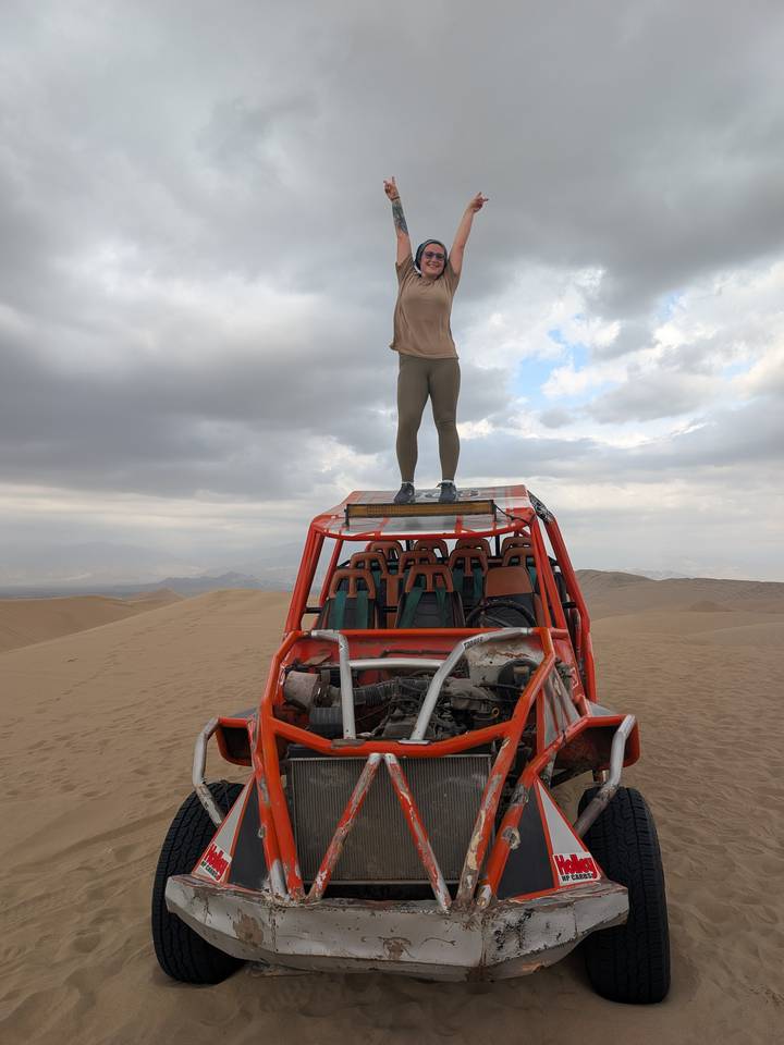 Traveller standing triumphantly on top of an open dune buggy in a vast sand-dune desert.