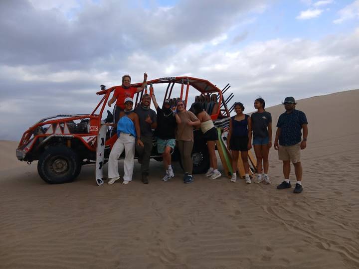 Group of travellers posing with a dune buggy and sandboards on a desert plain under cloudy sky.