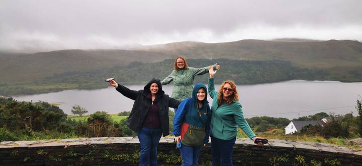 Four friends in rain jackets posing happily before a misty lake and rolling Irish hills.