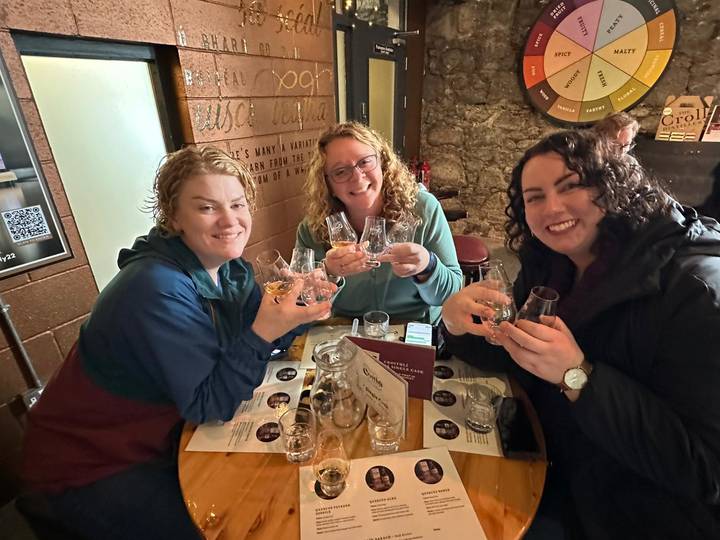 Three women clinking whisky tasting glasses inside a cosy Irish pub.