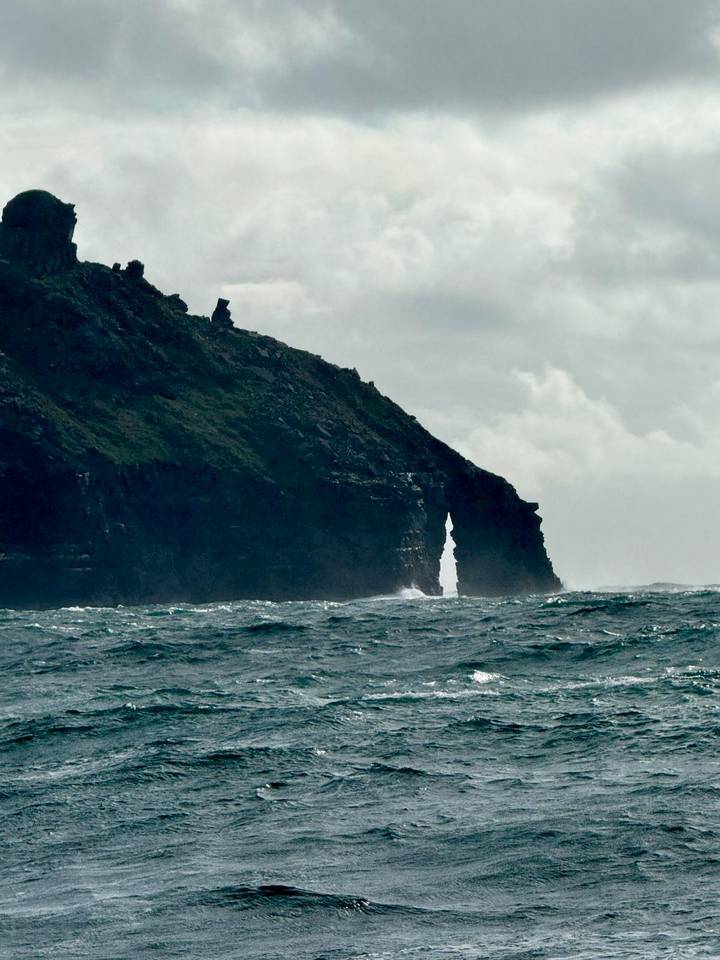Dramatic sea stack with a narrow arch battered by rough Atlantic waves under cloudy skies.