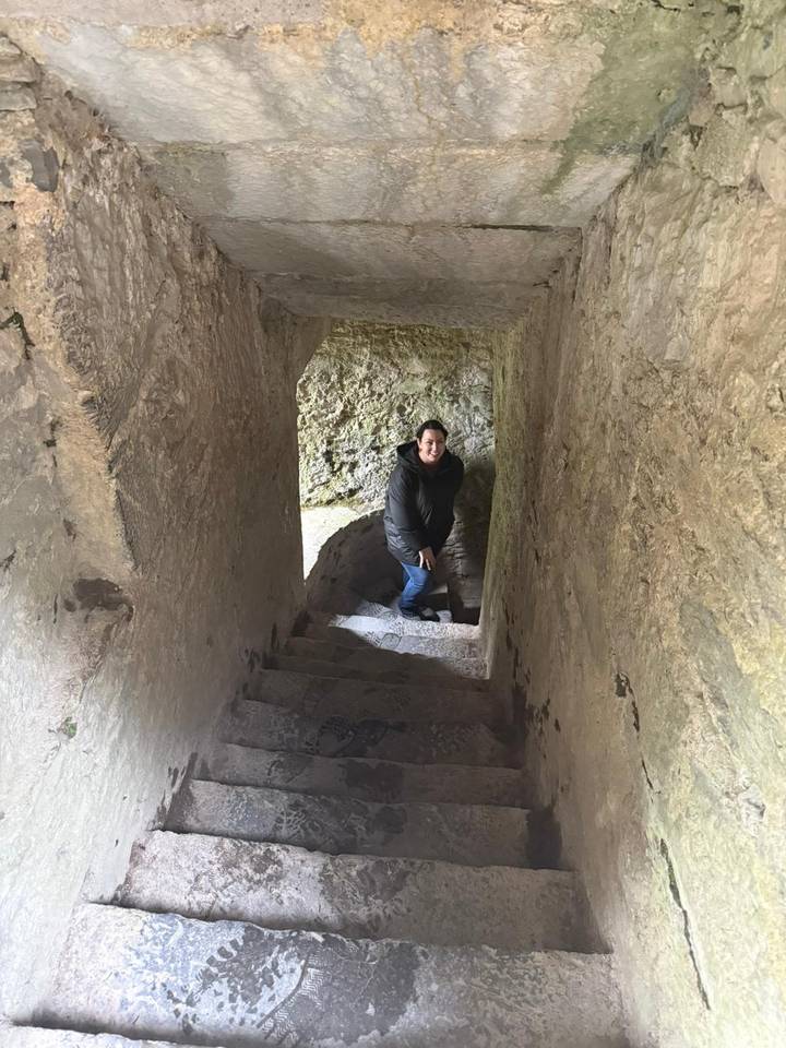Traveller smiling midway up a narrow stone stairwell inside an old castle tower.