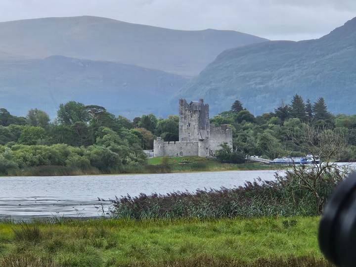 Stone castle stands on a wooded lakeside peninsula backed by misty mountains.