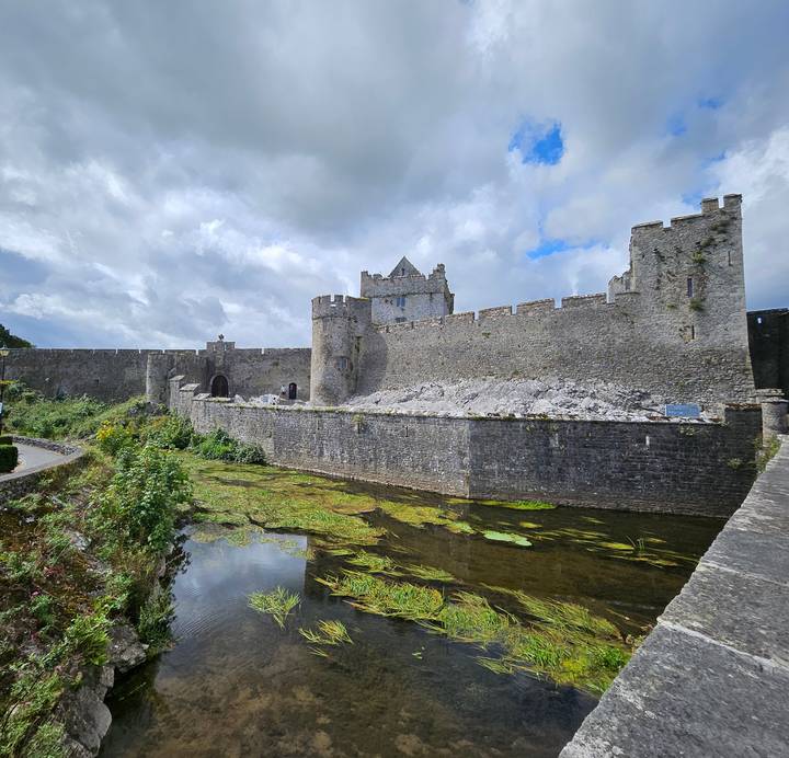 Medieval stone fortress with moat and rugged walls under a partly cloudy sky.