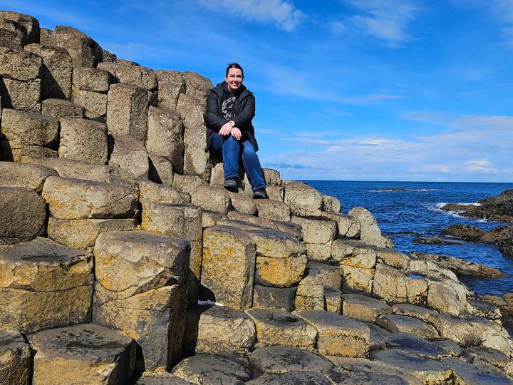 Traveller seated on iconic basalt columns beside deep blue sea at the Giant's Causeway.