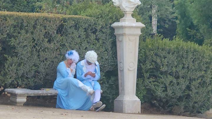 Two costumed performers in 18th-century attire sit on a bench in a manicured garden.