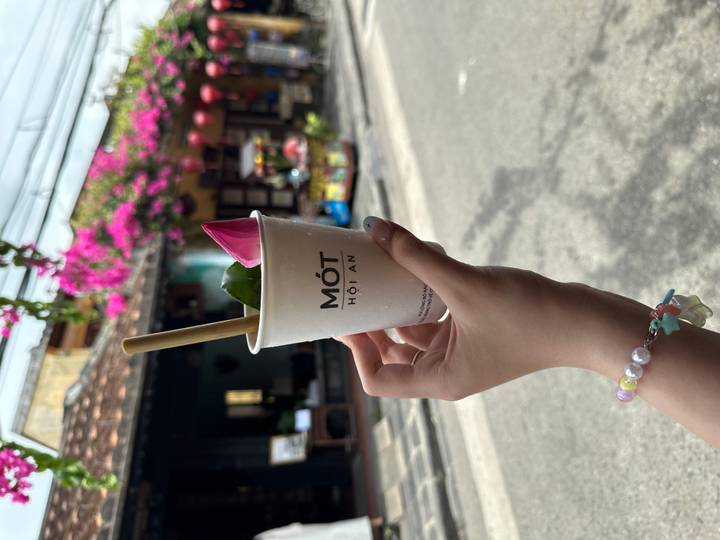 Hand holding a Mót Hoi An drink with bougainvillea and lantern-adorned street in background.