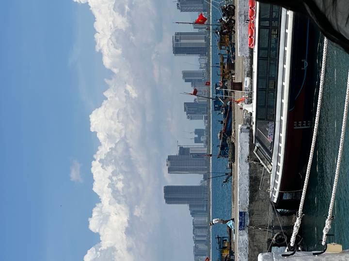 Harbor with speedboat, fishing vessels and modern high-rise skyline under a bright sky.