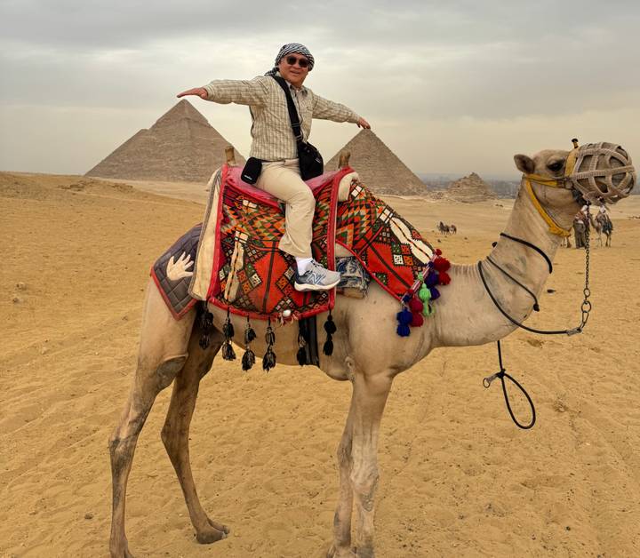 Traveler seated on a decorated camel pointing toward the pyramids on a sandy plateau in Giza