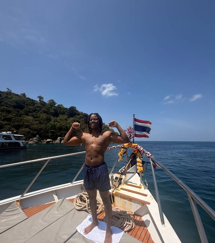 Traveller flexes muscles on a boat deck decorated with garlands and Thai flag against open sea.