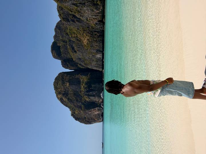 Man gazes over crystal-clear lagoon and dark limestone cliffs from pristine white sand.