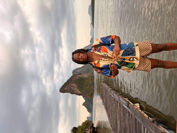 Man poses on a wooden pier with a dramatic limestone pinnacle and overcast sky beyond calm water.
