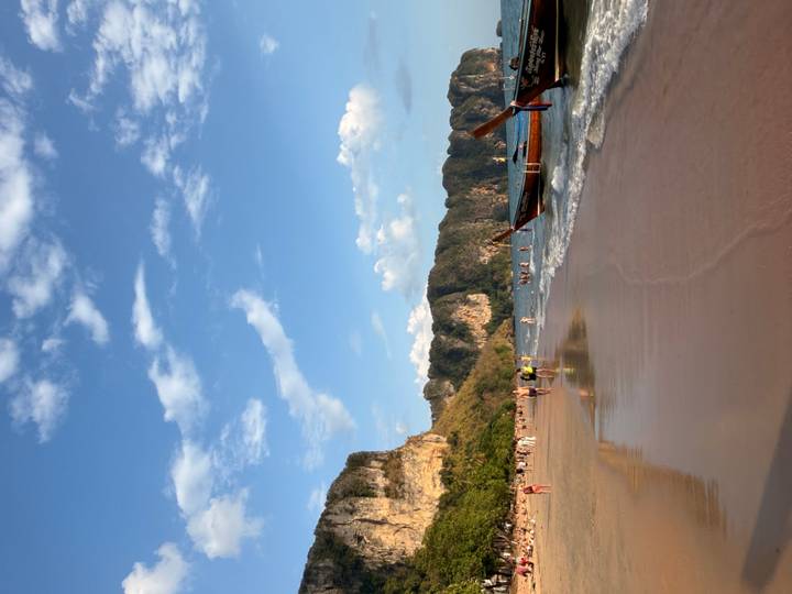 Sandy beach at sunset with long-tail boat ashore and people wading in gentle waves under blue sky.