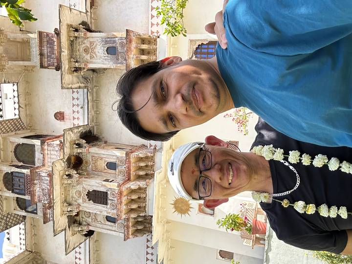 Two men take a close-up selfie in front of an ornate, weathered palace facade on a sunny day.