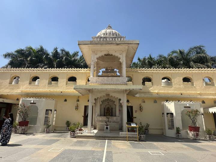 Elegant yellow-stone temple courtyard with carved arches, domed pavilion and palm trees under a clear sky.