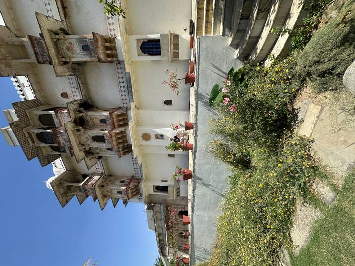 White heritage palace with intricate balconies overlooks a garden of flowering shrubs under blue skies.