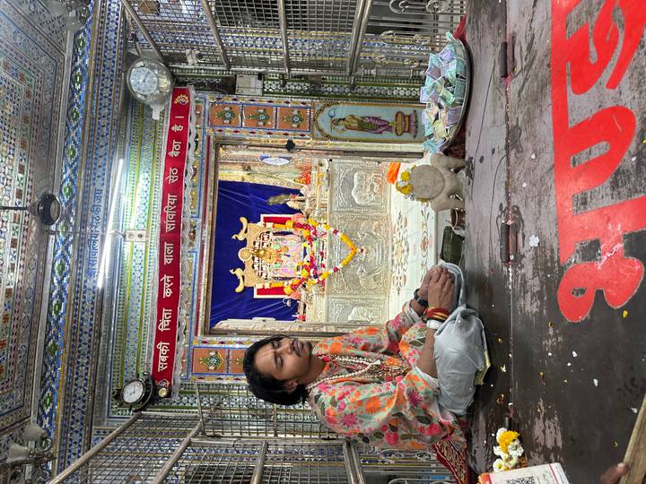 Young priest or devotee sits cross-legged inside a richly decorated Hindu shrine before a deity statue.