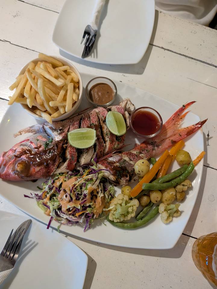 Plated whole grilled fish served with fries, vegetables, lime wedges and dipping sauces on a white dish.