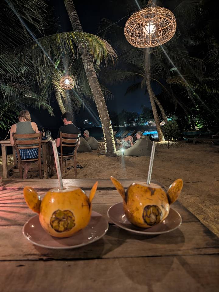 Nighttime beach restaurant with guests dining on sand under palm trees; coconut cocktails in foreground.