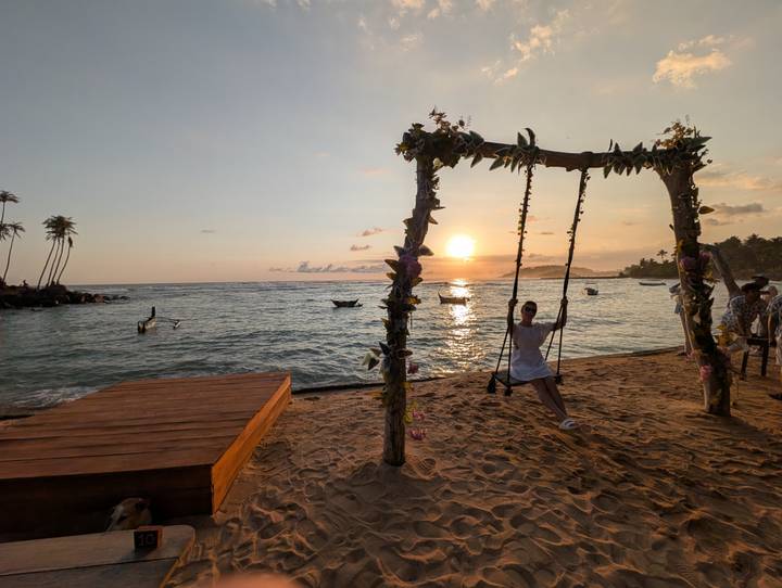 Person swings from a decorated wooden frame at sunset on a sandy tropical beach with calm sea.