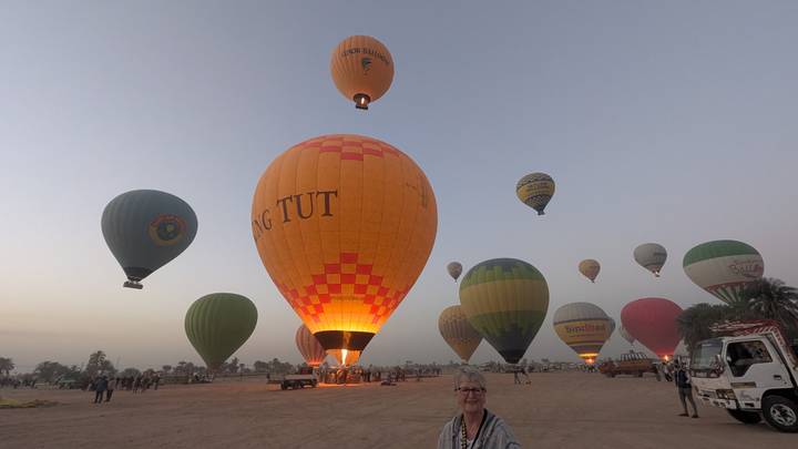A field of colorful hot-air balloons inflates at dawn with a smiling traveller in the foreground.