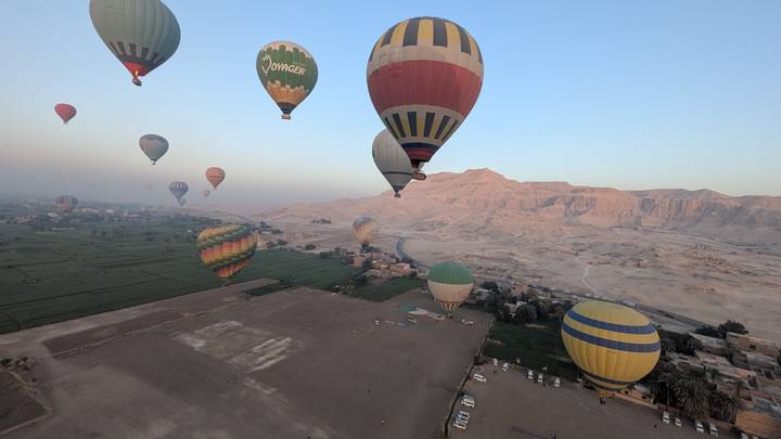 Aerial view of dozens of hot-air balloons drifting over green fields and desert cliffs near Luxor at sunrise.