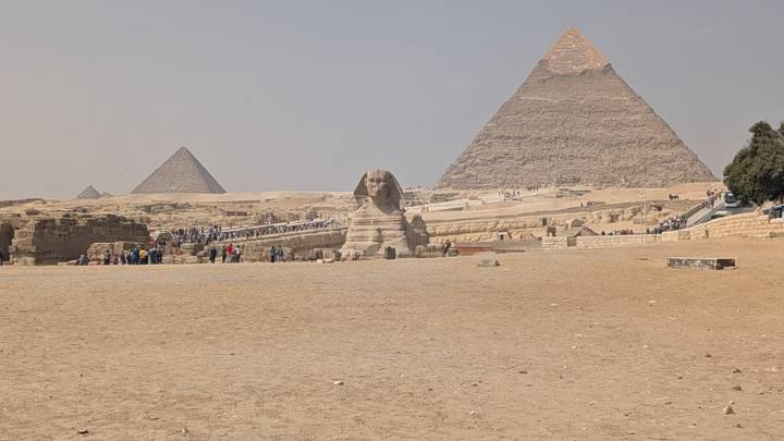 The Great Sphinx sits before the towering pyramids under a hazy sky with tourists scattered in the distance.