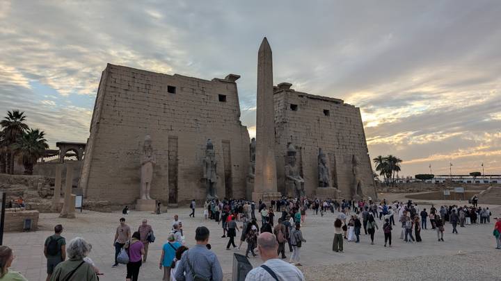 Crowds gather at the monumental entrance pylons and obelisk of Luxor Temple under a dramatic evening sky.