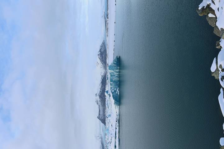 Serene glacial lagoon with a blue iceberg floating before snow-covered mountains under a pale sky.