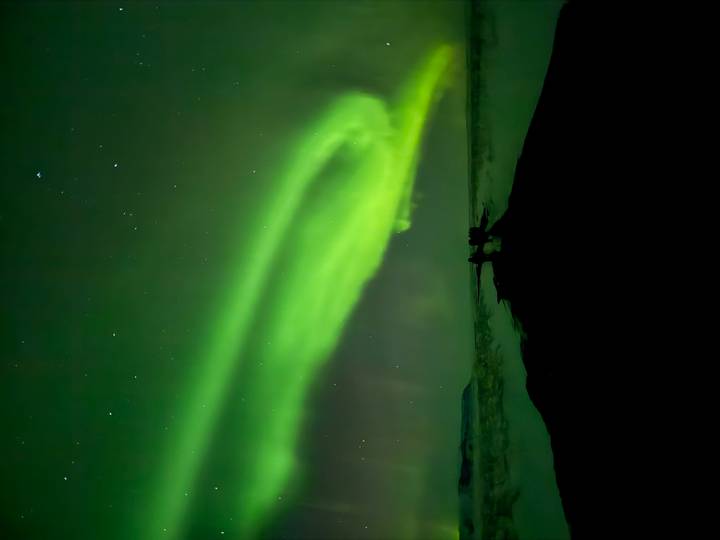 Vibrant green aurora dancing across the night sky above a dark snowy landscape with tiny silhouetted onlookers.