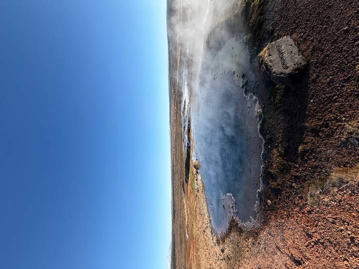 Steaming geothermal pool amid a barren plain under a clear blue sky.
